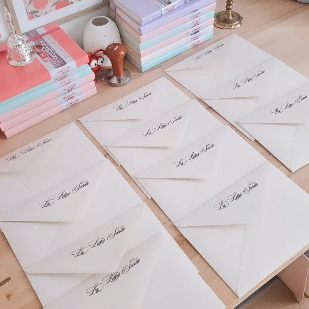 Rangées d'enveloppes crème calligraphiées La Lettre Secrète disposées sur un bureau en bois, entourées de livres pastel et d'un tampon encreur