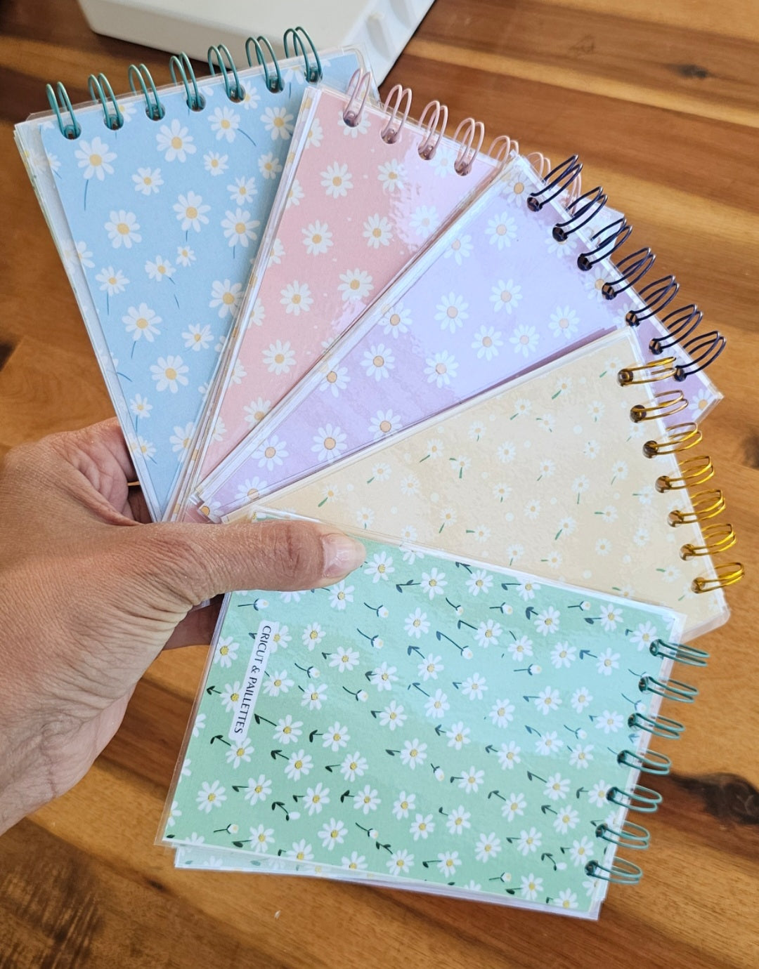 Main tenant plusieurs carnets printaniers faits main avec motifs de marguerites en couleurs pastel, reliés à spirale, sur une table en bois avec outil de reliure en arrière-plan